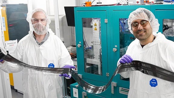 Zwei Männer in weißen Laborkitteln halten eine mehrere Meter lange, schmale und sehr dünne Solarfolie in die Kamera.