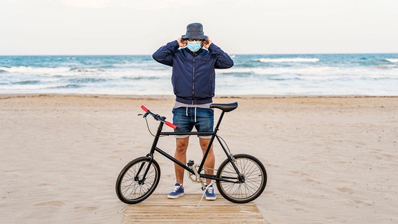 Frontale Ansicht: Mann mit Angelhut setzt sich am Strand OP-Maske auf, vor ihm Fahrrad mit kleinen Rädern, Hintergrund Meer und Horizont