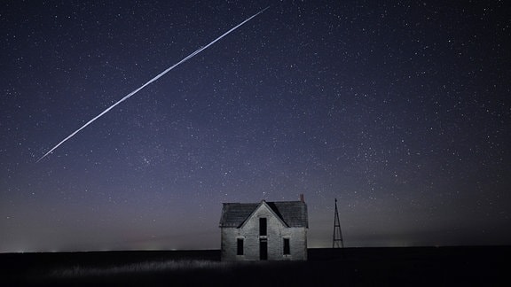 Langzeitbelichtet bei Nacht: Einsames altes Haus vor großer Freifläche, darüber Sternenhimmel und lange Lichtspur durch Starlink-Satelliten (ähnlich wie Sternschnuppe)