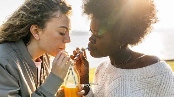 Zwei Frauen mit weißer und schwarzer Hautfarbe trinken aus einer Flasche mit Orangensaft mit zwei gebogenen Papierstrohhalmen, die sich überkreuzen. Seitenansicht mit Gegenlicht der Sonne