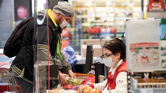 Ein Mann steht an einer Kasse im Supermarkt. Käufer und Verkäuferin tragen Mundschutz.