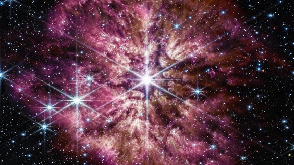 Der leuchtende, heiße Stern Wolf-Rayet 124 (WR 124) ist im Zentrum des zusammengesetzten Bildes des James Webb Weltraumteleskops zu sehen, das Licht aus dem nahen und mittleren Infrarot kombiniert.   Hintergrundsterne und Hintergrundgalaxien bevölkern das Sichtfeld und blicken durch den Gas- und Staubnebel, der von dem alternden massereichen Stern ausgestoßen wurde und sich über 10 Lichtjahre im Weltraum erstreckt. An der Struktur des Nebels lässt sich die Geschichte der vergangenen Massenauswürfe des Sterns ablesen. Der Nebel besteht nicht aus glatten Schalen, sondern aus zufälligen, asymmetrischen Auswürfen. Helle Gas- und Staubklumpen erscheinen wie Kaulquappen, die auf den Stern zuschwimmen, mit Schwänzen, die hinter ihnen herausströmen und vom Sternwind zurückgeblasen werden.