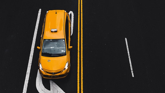 Draufsicht auf eine schwarze Straße mit gelben Mittellinien und weißen Markierungen. Darauf ein gelbes Taxi.