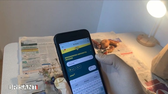Hand mit weißem Gummihandschuh hält Smartphone mit Resteretter-Webapp und trägt "Brötchen" ein. Unscharf im Hintergrund auf Zeitungspapier Lebensmittelreste vom Obst, Backwaren.