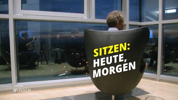 Drehsessel umgedreht, vor hohen Fenstern in Hochhaus, darauf Schrift "Sitzen: heute, morgen"