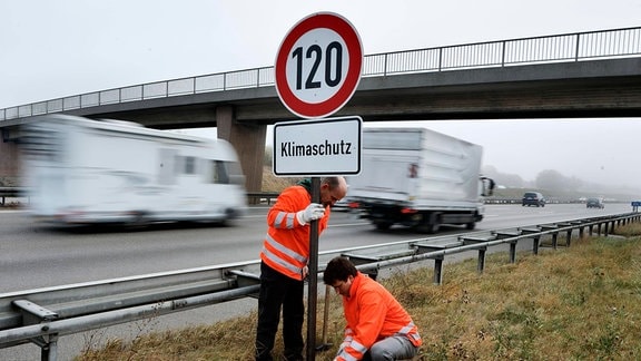 Zwei Männer mit organgefarbener Warnjacke stellen ein Tempo-120-Schild mit Zusatzinfo "Klimaschutz" an einer befahrenen Autobahn auf.