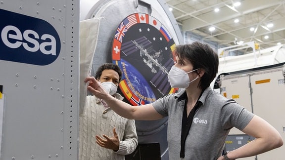 Frau mit kurzen dunklen Haaren und FFP2-Maske. Daneben mann mit dunklen, kurzen, lockigen Haaren und FFP2-Maske. ISS-Trainingsumgebung im HIntergrund, Esa-Logo im Vordergrund.