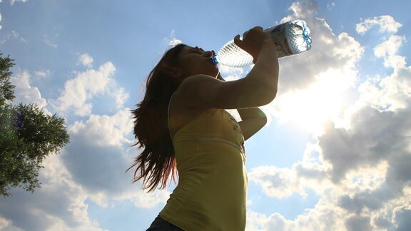 Frau mit dunklen, im Wind wehenden Haaren trinkt aus einer Wasserflasche, Foto aus Froschperspektive