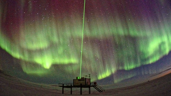 Dunkler Himmel mit grünen Schleiern, Polarlichter, im Vordergrund kleiner Forschungscontainer auf Gestell mit grüner Laser-Säule nach oben