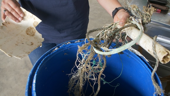 Eine Hand hält verschiedene Plastikteile sowie Nylonseile und Fischernetze über einer blauen Plastiktonne.