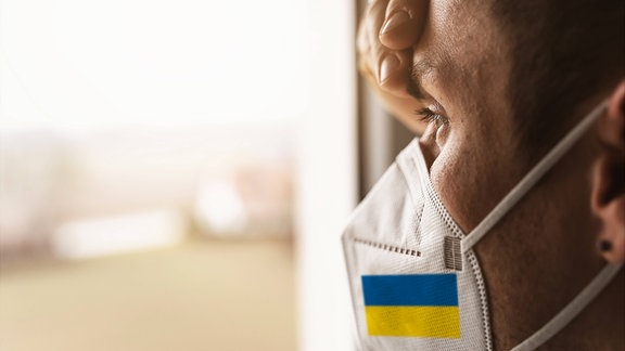 Mann an Fenster trägt medizinische Maske in den Farben der ukrainischen Flagge