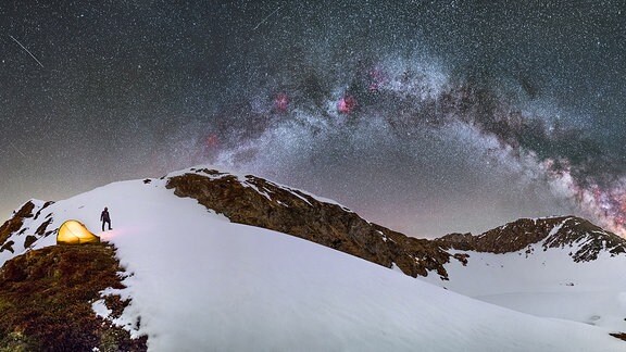 Nachthimmel mit Sternen vor verschneitem Berg, links ein Mensch und ein Zelt 