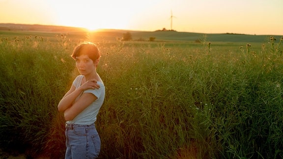 Eine junge Frau steht in der Abendsonne vor einem Feld, im Hintergrund ist ein Windrad zu sehen.
