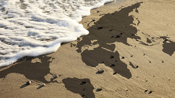 Die Weltkarte im Strandsand und eine Welle bricht am Strand.