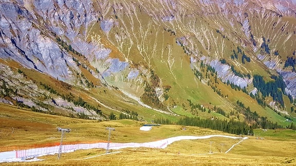 Eine Berglandschaft mit zerklüfteten Felsen. Über die grüne Wiese zieht sich eine weiß ausgelegte Kunstfaser-Piste 