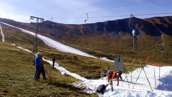 Ein Berghang, mit grüner Wiese. Darauf zwei weiße Streifen, ein Skilift.   