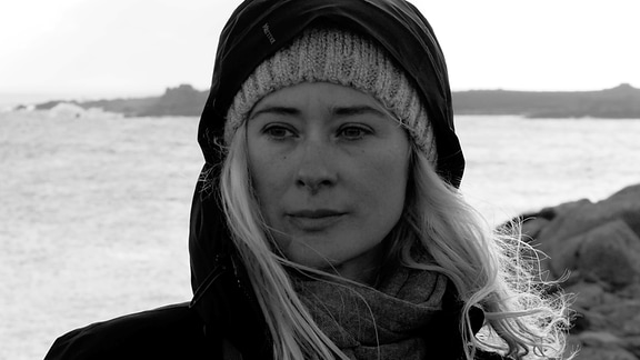 Auf dem schwarz-weiß Foto ist eine junge Frau mit langen blonden Haaren und Winterkleidung vor einer Küstenlandschaft zu sehen.