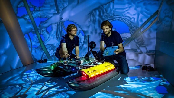 Zwei Männer hocken neben einem Spezialroboter und arbeiten an ihm.