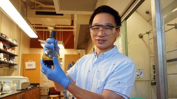 Ein junger Forscher ostasiatischer Abstammung mit schwarzem, kurzen Haar und einem blauen Hemd steht in einem Labor und hält ein Reagenzglas mit einer Flüssigkeit in die Kamera.  