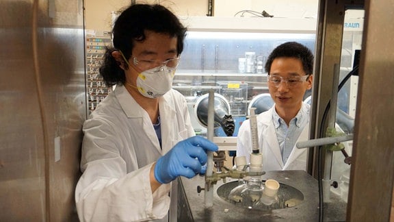 Zwei Forschende - eine Person mit langen schwarzen Haaren und ein Mann mit kurzen schwarzen Haaren - stehen mit Laborkitteln bekleidet in einem Labor und begutachten ein technisches Gerät. 