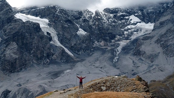 Herrliches Bergmassiv mit Gletscherzungen, im Vordergrund wnadernde Person mit ausgestreckten Armen und Wanderstöcken.