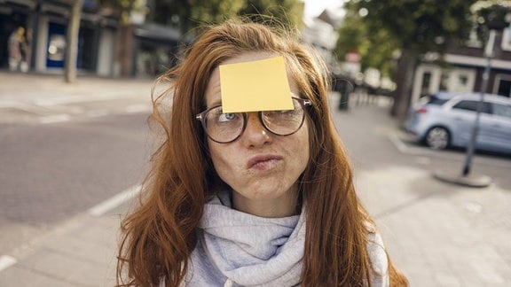 Junge Frau mit runder Brille und roten Haaren und leeren gelben Klebezettel auf der Stirn schaut verdutzt, Hintergrund unscharf Straße, Bäume, Auto
