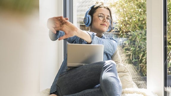 Junge Frau mit Kopfhörern und runder, drahtiger Brille sitzt am Fenster mit Laptop, schaut von Tablet glücklich weg und streckt Arme mit verschränkten Händen nach vorn.