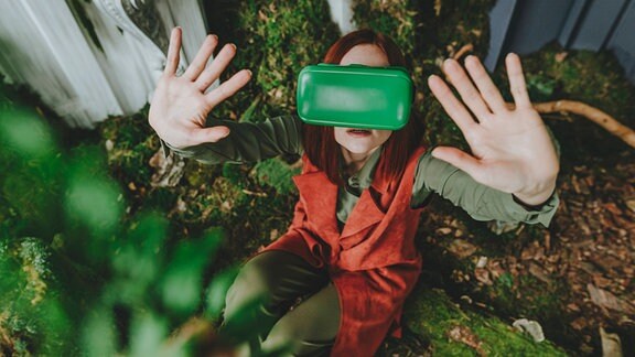 Von oben: Frau trägt markante grüne VR-Brille mit Blick nach oben in Kamera, streckt Arme entgegen. Befindet sich in Wohnung umgeben mit Pflanzen und Holz-Mulch-artigem Boden.