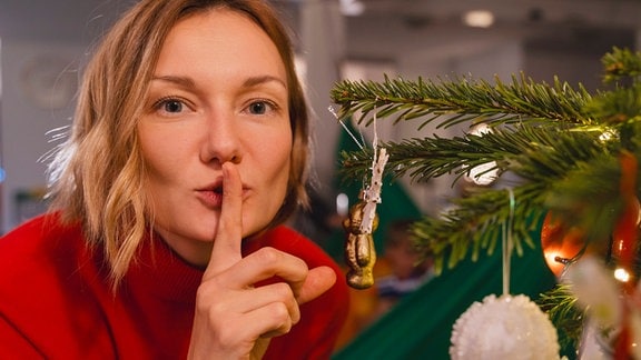 Eine Frau beugt sich neben einem Weihnachtsbaum zur Kamera und hält ihren Finger vor den Mund.