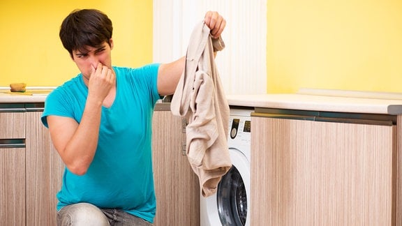 Junger Mann hockt vor einer Waschmaschine und hebt ein Wäschestück an, während er sich gleichzeitig die Nase zuhält.