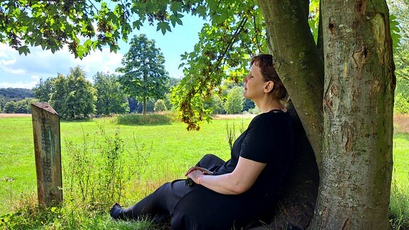 Eine junge Frau sitzt mit geschlossenen Augen an einen Baum gelehnt auf einer Waldwiese.