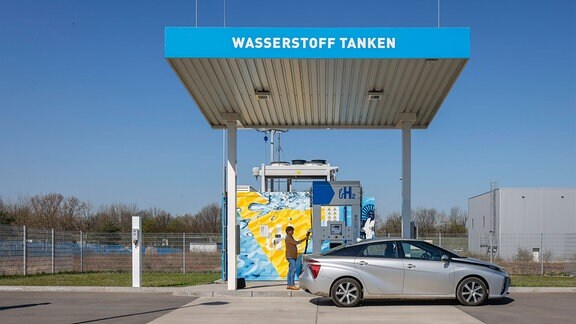 Kleines Tankstellengebäude: Auto steht davor, Person tankt Wasserstoff. Aufschrift auf Tankstellendach "Wasserstoff tanken". 