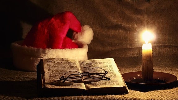 Eine Brille auf einem aufgeschlagenen Buch, eine brennende Kerze und eine Weihnachtsmannmütze