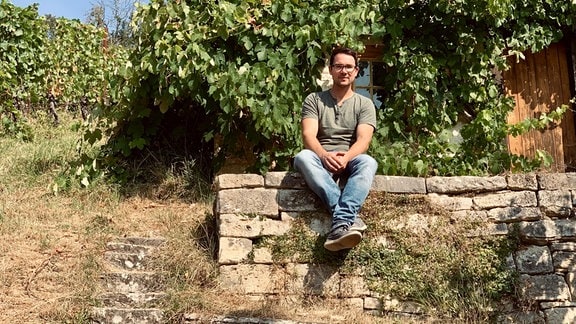 Junger Mann sitzt auf Steinmauer in Weingut vor kleinem Häuschen, im Hintergrund Weinreben, trockenes, warmes Wetter, Steinstufen