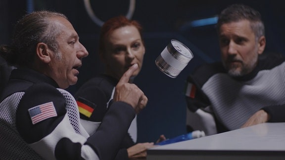 Drei Raumfahrende mit Länderflagge auf Arm an Tisch in Schwerelosigkeit, Mann zeigt auf schwebende Konservendose