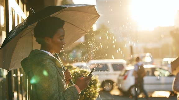 Seitenansicht: Frau steht unter Regenschirm auf den Regen spritzt in urbanem Umfeld, Gegenlicht durch Sonne, Frau schaut aufs Handy