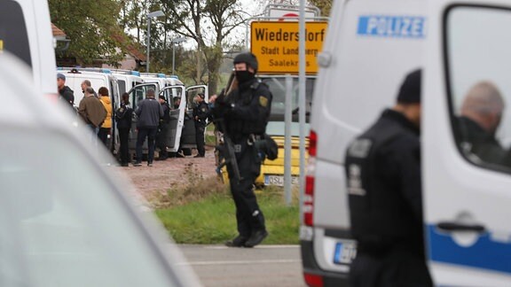 Polizeieinsatz: SEK-Polizist vor Ortsschild Wiedersdorf, unscharf im Vordergrund Polizeitransporter, weitere Polizeitransporter, Polizistinnen und Polizisten und andere Personen im Vorder- und Hintergrund.