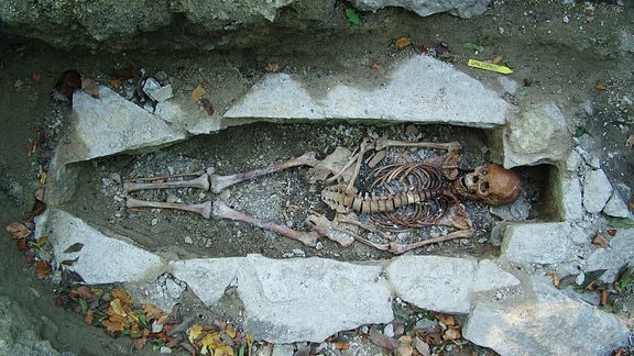 Das Skelett einer Frau liegt in einem offenen Erdgrab umrandet von Steinen.