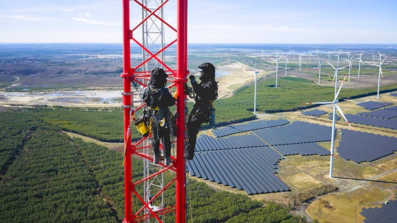 Zwei Kletterer auf einem roten Stahlturm. Im Hintergrund ein Solar- und Windpark.