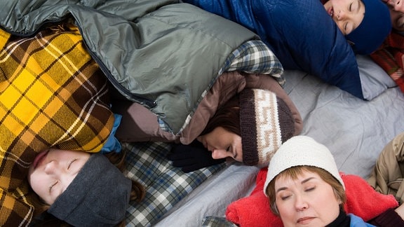 Blick von oben: Kinder und Erwachsene liegen kreuz und quer in Zelt im Schlafsack mit Mützen und schlafen.