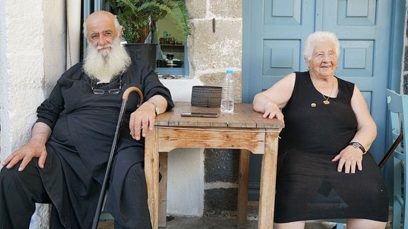 Porträt eines älteren Herren und einer älteren Dame, die vor einem Haus sitzen.