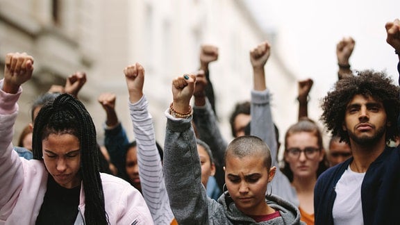 Gruppe mit Frauen und Männern verschiedener ethnischer Abstammungen frontal in einem Protestmarsch mit erhobenen Fäusten und ernsten, teils wütenden Blick, teilweise nach unten