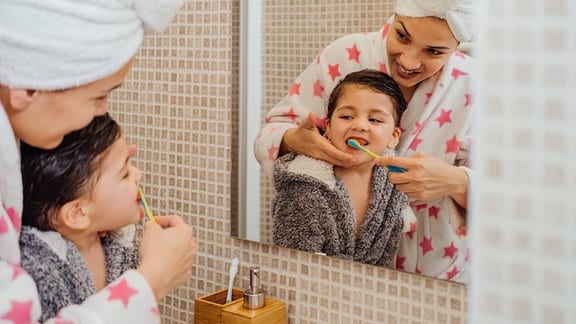 Blick auf Spiegelbild: Mutter mit Handtuch um Kopf und Bademantel putze die Zähne eines lachenden kleinen Jungen