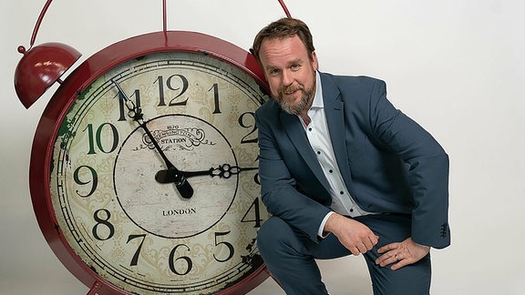 Ein Mann einem Anzug sitzt vor einer großen Uhr.