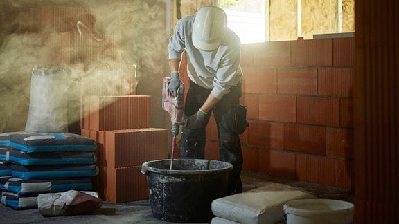 Person mit Bauhelm nach unten gebeugt, mischt Zement in einem Bottich auf Baustelle in einem Haus-Rohbau, Sonnenstrahlen treten ein und sorgen für stimmungsvolles Gegenlicht